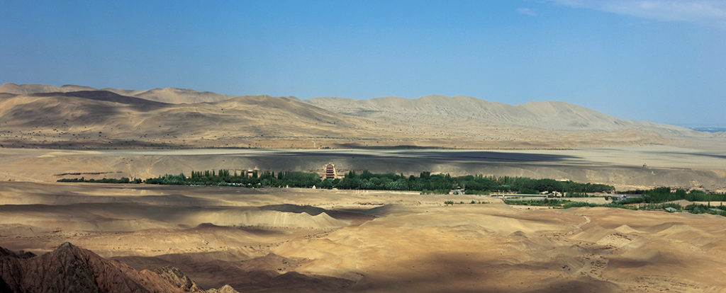 The cave temples of Mogao, carved into the cliff face along the Daquan River, are shaded by poplar trees planted in the 20th century and surrounded by austere desert. The nine- story temple can be seen at the center. Beyond the plateau above the cliff rise the Mingsha Shan—the Dunes of the Singing Sands. Photo: Sun Zhijun, ©Dunhuang Academy